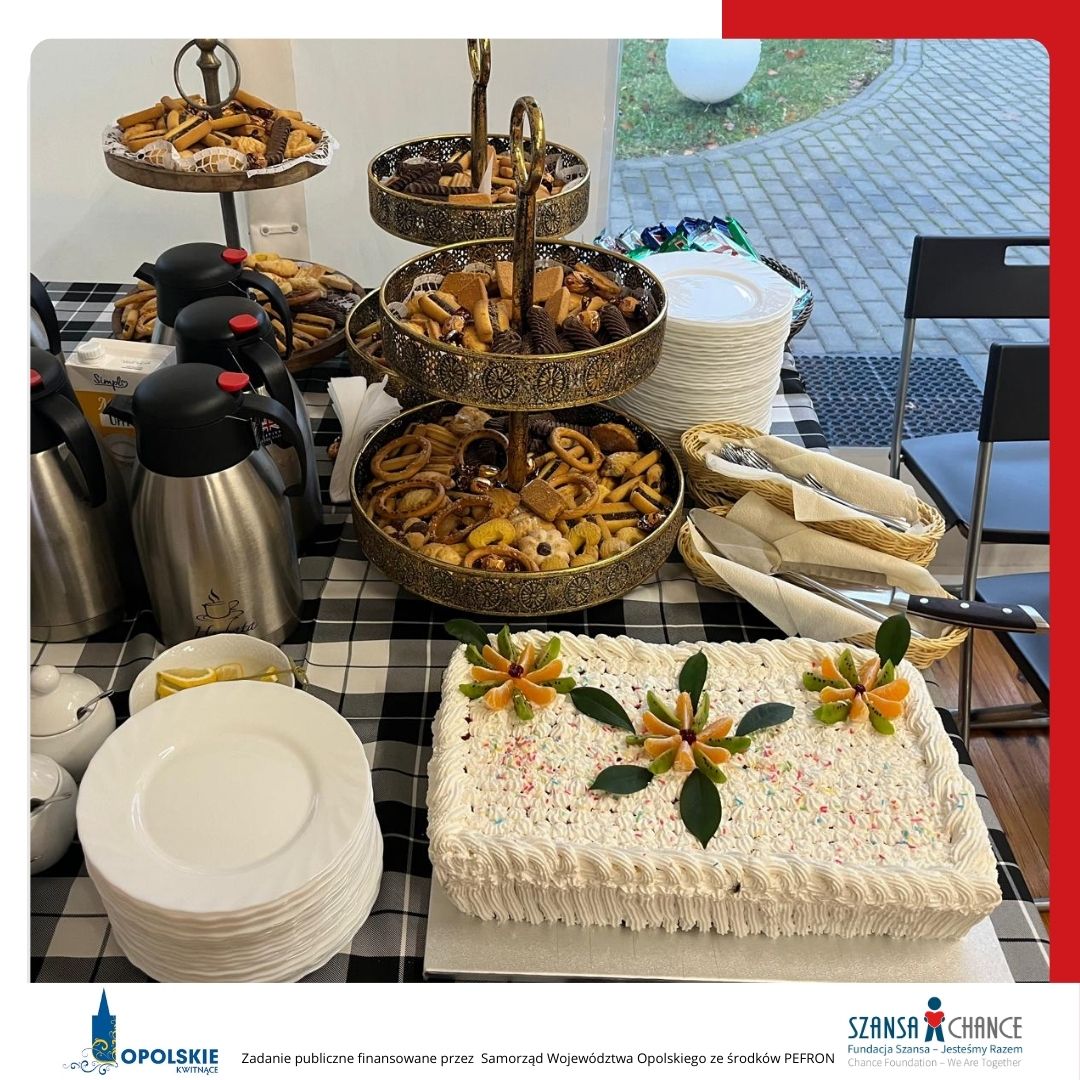 Stół z przekąskami i deserami. Na pierwszym planie duży prostokątny tort udekorowany owocami i kremem. Obok znajdują się paterki z ciasteczkami, termosy z kawą i herbatą oraz stosy talerzy i sztućców. W dolnej części zdjęcia znajdują się logotypy: Opolskie Kwitnące oraz Fundacja Szansa- Jesteśmy Razem. Tekst informuje, że zadanie jest finansowane przez Samorząd Województwa Opolskiego ze środków PFRON.