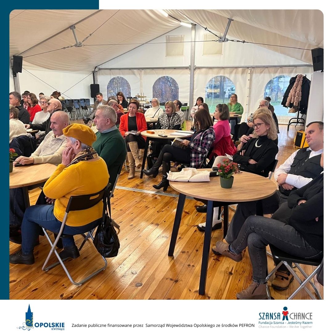Widok na uczestników wydarzenia siedzących w namiocie przy okrągłych stolikach. Ludzie uważnie słuchają, niektórzy robią notatki. Pomieszczenie jest jasne, z widocznymi dużymi oknami w tle. W dolnej części zdjęcia znajdują się logotypy: Opolskie Kwitnące oraz Fundacja Szansa- Jesteśmy Razem. Tekst informuje, że zadanie jest finansowane przez Samorząd Województwa Opolskiego ze środków PFRON.